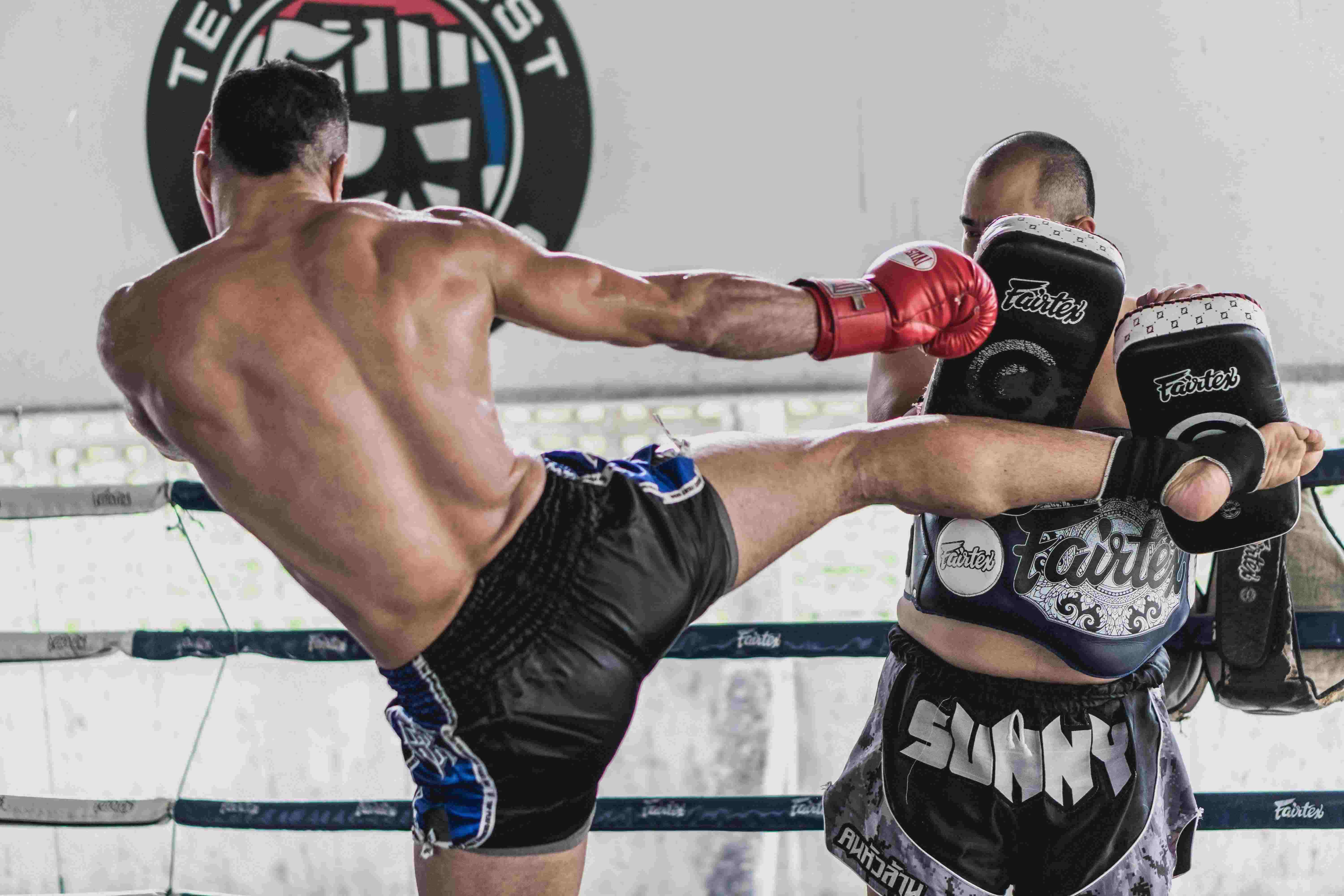 A Caucasion adult male with no shirt and black shorts practices muay thai by kicking mitts held by another person.